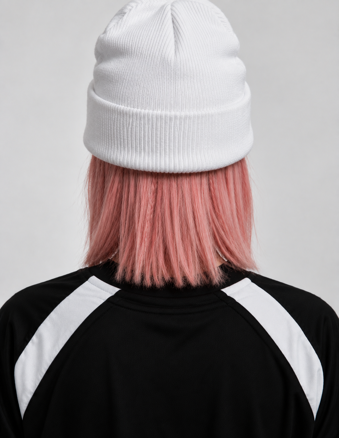 A person shown from the back wearing a white ribbed knit beanie, pink hair, and a black and white athletic top against a light gray studio background.